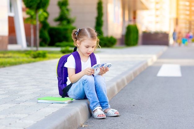 Een gelukkig kind, een schoolmeisje met een rugzak en rekeningen in haar handen, zit op de stoep bij de school en leert getallen te tellen, het concept is terug naar school