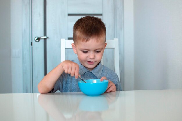 Een gelukkig kind eet een gezond heerlijk ontbijt. Portret van een kind aan het ontbijt. Een schattige gelukkige baby zit in de keuken en leert met een lepel. Selectieve aandacht.