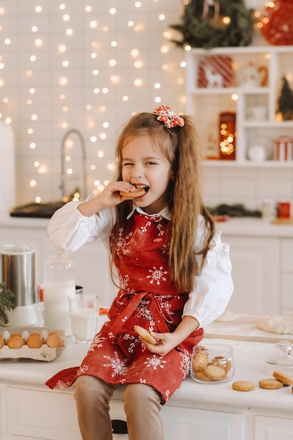 Een gelukkig klein meisje in de kerstkeuken zit op tafel met koekjes in haar handen en eet het op.