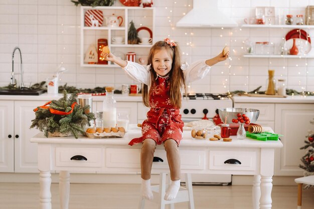 Een gelukkig klein meisje in de kerstkeuken zit op tafel met koekjes in haar handen.