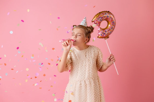 Een gelukkig klein meisje in een verjaardagspet houdt een donutvormige ballon op een roze achtergrond in de studio