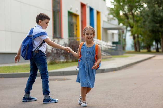 Een gelukkig klein meisje met een oranje rugzak en een broer met blauwe rugzakken houden elkaars hand vast, spelen en hebben plezier in de buurt van het schoolgebouw op een zonnige dag in de vroege herfst. Kopieer ruimte