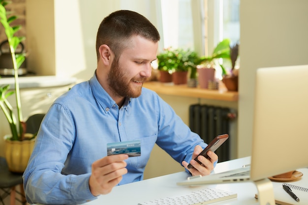 Een gelukkig man met een baard op zoek naar producten in online winkels op een smartphone en in zijn hand met een creditcard thuis