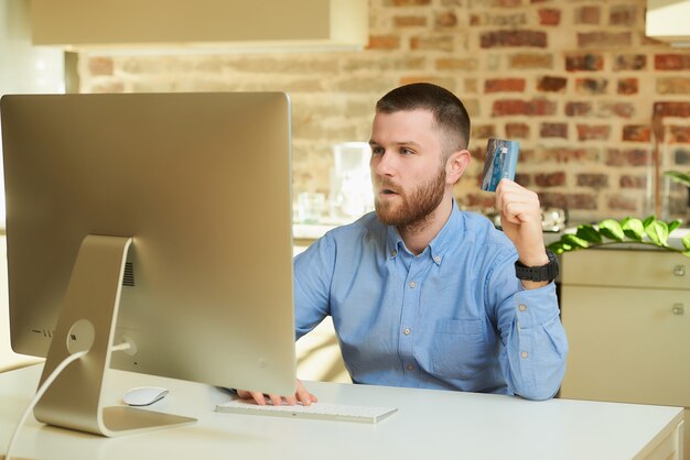 Een gelukkig man met een baard zit achter de computer verrast door de prijzen op een online winkel thuis