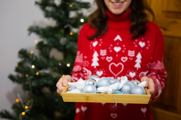 Een gelukkig meisje houdt een dienblad vast waarin veel mooie veelkleurige kerstversieringen zijn. Kerstboom decoratie proces. Nieuwjaar Kerstmis. 2022 jaar van de tijger.