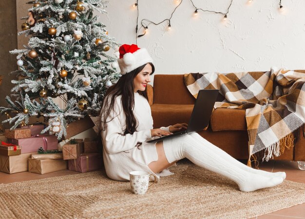 Een gelukkig meisje met een kerstmuts wenst haar vrienden prettige kerstdagen in een videochat op een laptop Een jonge vrouw ligt onder een kerstboom tussen geschenkdozen in een interieur