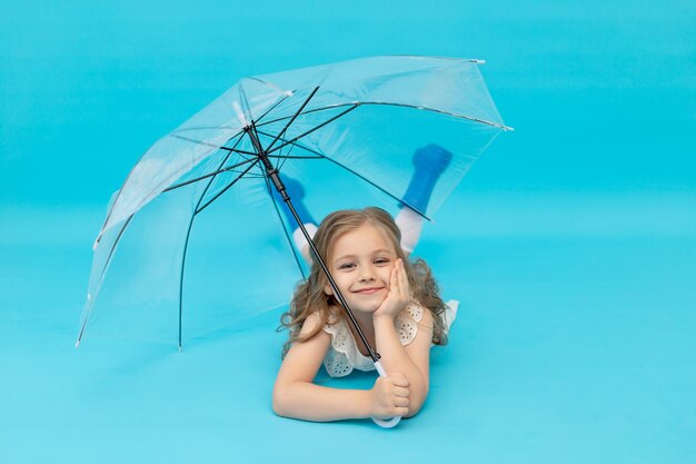 Een gelukkig schattig klein meisje in blauwe rubberen laarzen en een katoenen witte jurk met een paraplu liggend op een blauwe achtergrond in de studio lachend, glimlachend en gek rond, een plek voor tekst