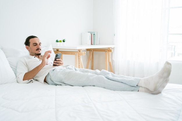 Een gelukkige, aantrekkelijke, bebaarde blanke man die 's ochtends op het bed ligt met vape in een appartement met behulp van een smartphone die sociale media controleert, winkelt en chat met vrienden Afbeelding met kopieerruimte