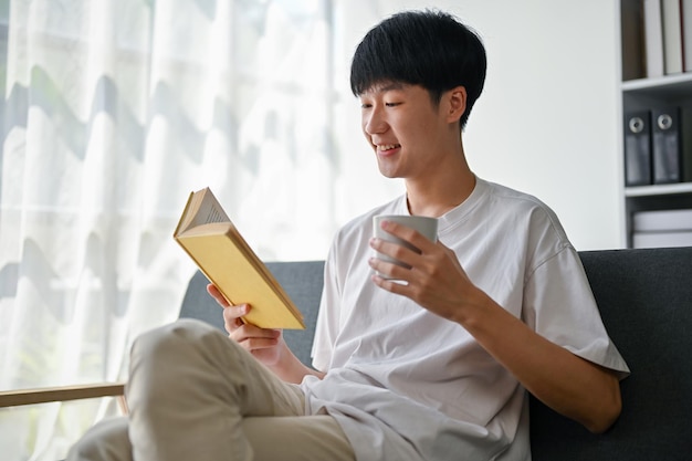 Een gelukkige Aziatische man leest een boek terwijl hij geniet van zijn ochtendkoffie op een sofa in de woonkamer