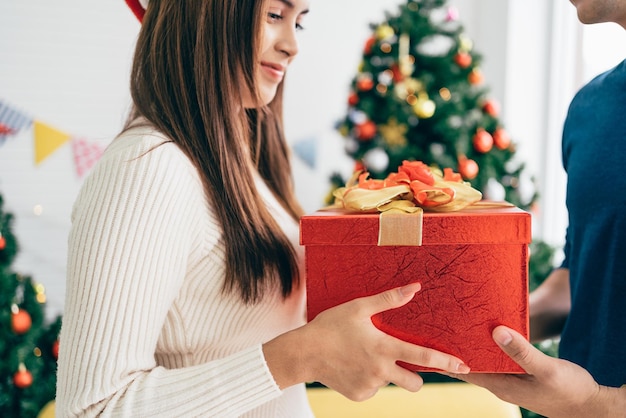 Een gelukkige Aziatische man verrast zijn vriendin met kerstcadeautjes thuis met een kerstboom op de achtergrondafbeelding met kopieerruimte