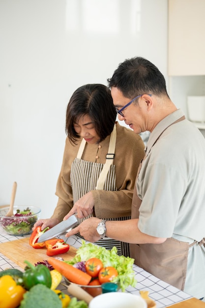 Een gelukkige aziatische vrouw hakken een zoete peper terwijl haar man haar helpt in de keuken