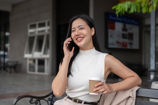 Een gelukkige Aziatische vrouw praat aan de telefoon en drinkt koffie terwijl ze op een bankje in de stad rust