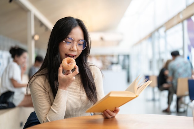 Een gelukkige Aziatische vrouw zit in een koffieshop een boek te lezen en te genieten van donuts