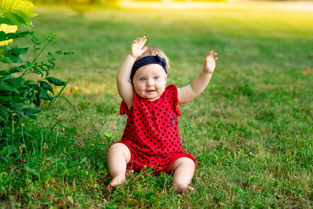 Een gelukkige baby in de zomer op het groene gras in een rode bodysuit stak zijn handen omhoog van vreugde in de ondergaande zon, ruimte voor tekst