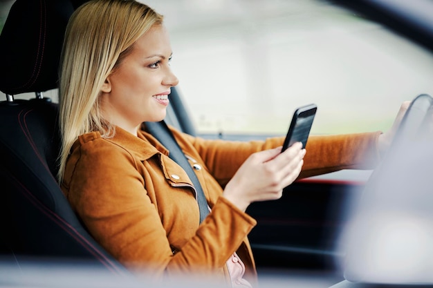 Een gelukkige blonde vrouw zit in haar auto en gebruikt haar telefoon