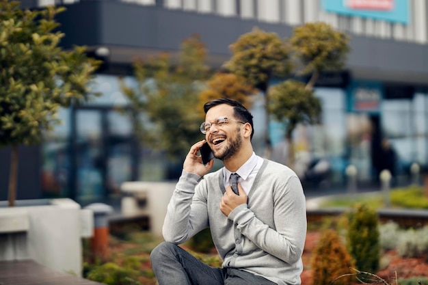 Een gelukkige elegante man zit buiten en lacht terwijl hij een telefoongesprek voert