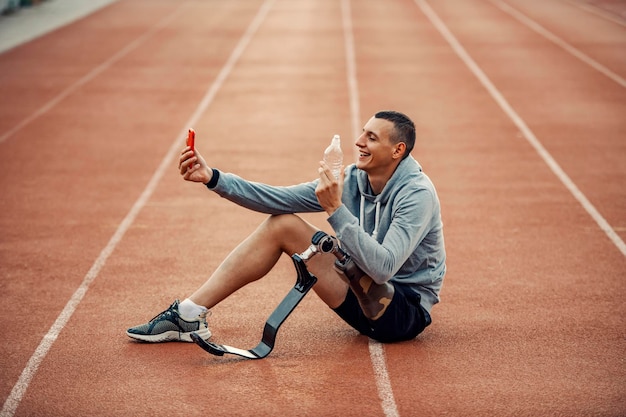 Een gelukkige gehandicapte sporter met een beenprothese die selfie neemt met een fles water in het stadion