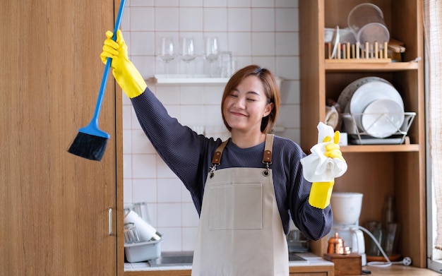 Een gelukkige huisvrouw met een beschermende handschoen die een bezem vasthoudt en danst terwijl ze thuis in de keuken schoonmaakt