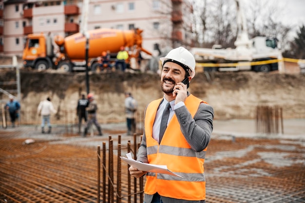 Een gelukkige ingenieur met papierwerk in zijn handen staat ter plaatse en heeft een telefoontje