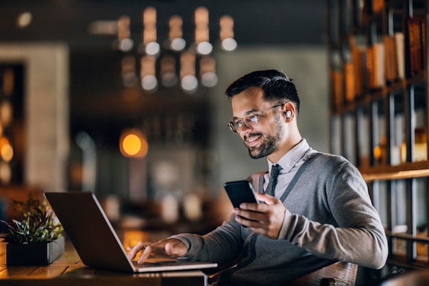 Een gelukkige jonge elegante ondernemer zit in een restaurant en typt een rapport terwijl hij de telefoon gebruikt