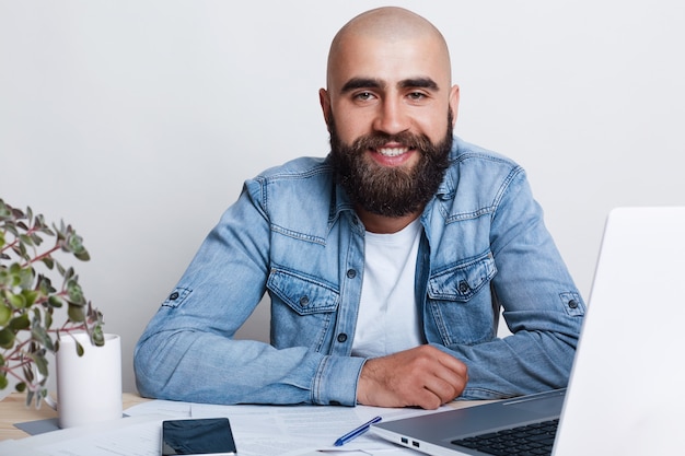 Een gelukkige jonge kale zakenman die dikke donkere baard gekleed in het overhemd van Jean siiting bij de lijst in zijn bureau met laptop heeft