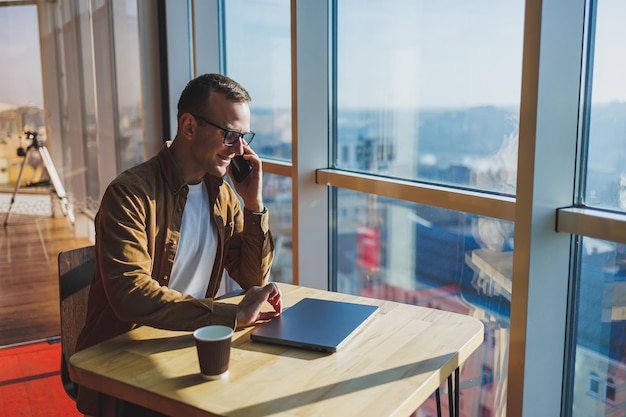 Een gelukkige jonge man met een bril en vrijetijdskleding praat aan de telefoon terwijl hij op een laptop werkt vanuit een gezellige werkruimte Een succesvolle freelancer werkt op afstand