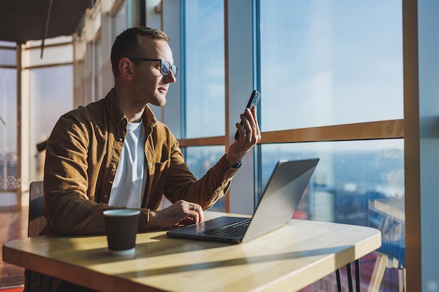 Een gelukkige jonge man met een bril en vrijetijdskleding praat aan de telefoon terwijl hij op een laptop werkt vanuit een gezellige werkruimte Een succesvolle freelancer werkt op afstand