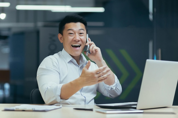 Een gelukkige jonge man zit op kantoor aan de tafel, hij praat aan de telefoon die hij goed heeft ontvangen