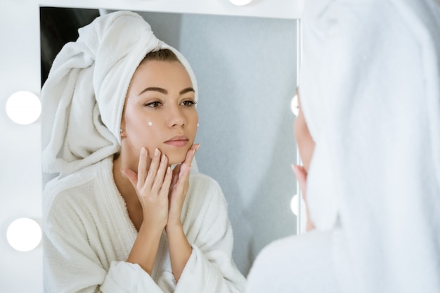 Een gelukkige jonge vrouw in een handdoek voor een spiegel past room toe op haar gezicht, een concept van huidverzorging thuis