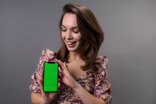 Een gelukkige jonge vrouw kijkt naar het smartphonescherm met een chromakey op een grijze achtergrond Emotioneel model in stijlvolle kleding