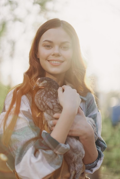 Een gelukkige jonge vrouw lacht naar de camera en houdt een jonge kip vast die eieren legt voor haar boerderij in het zonlicht