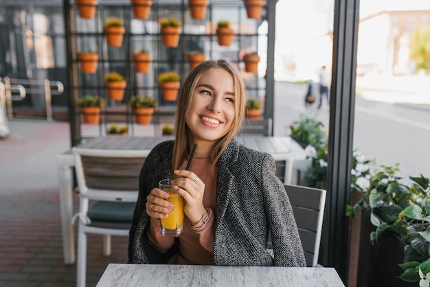 Een gelukkige jonge vrouw zit aan een tafel in een zomercafé en houdt een glas mangosap in haar hand