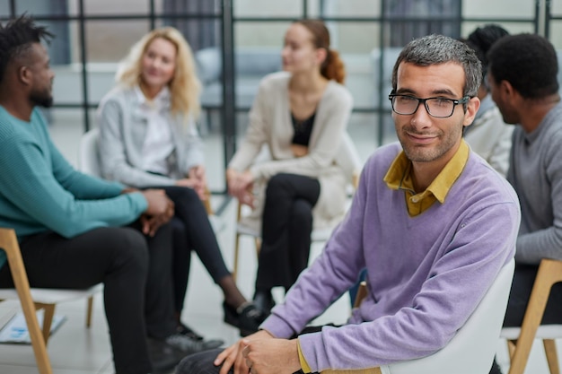 Een gelukkige jonge zakenman in een paarse trui zit tegen de achtergrond van zijn collega's
