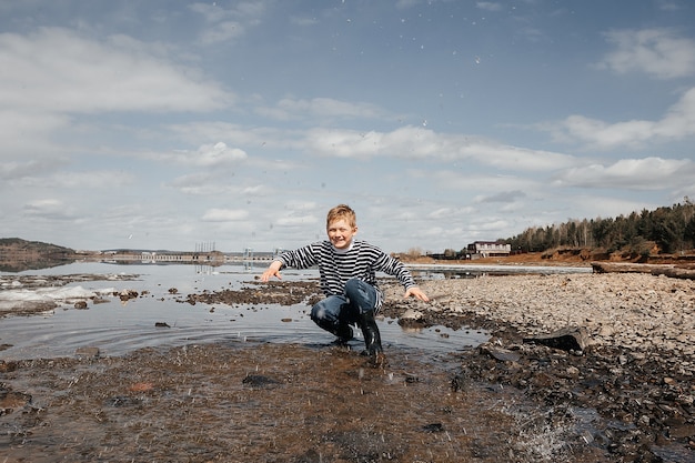 Foto een gelukkige jongen in een gestreept vest en rubberen laarzen aan de oever van de rivier spettert vrolijk water. buitenrecreatie.