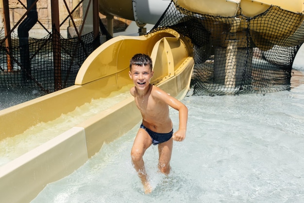 Een gelukkige jongen van zeven jaar oud glimlacht en toont een klas tegen de achtergrond van dia's in een waterpark Prettige vakantievakantie Zomervakantie en toerisme