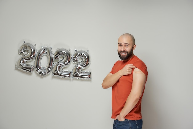 Een gelukkige kale man met een baard toont een medische pleister op zijn schouder na vaccinatie in de buurt van verijdelde zilveren ballonnen in de vorm van 2022.