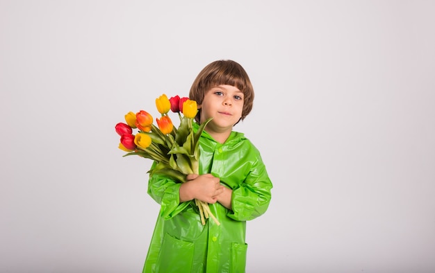 Een gelukkige kleine jongen in een groene regenjas houdt een boeket kleurrijke tulpen vast op een witte achtergrond met een plek voor tekst