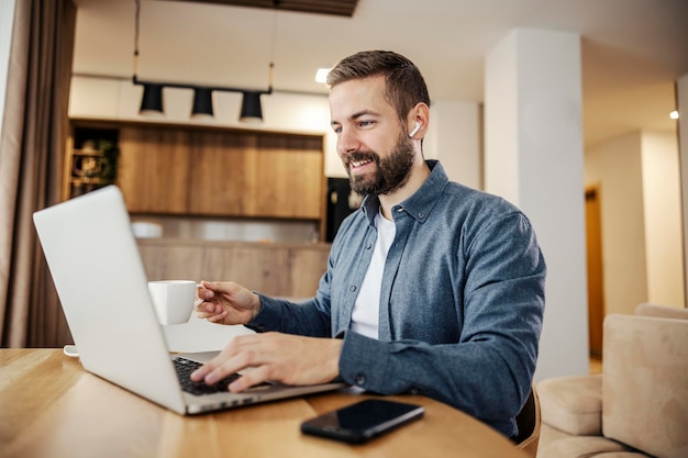 Een gelukkige man drinkt zijn ochtendkoffie en voltooit zijn werk op afstand op een laptop vanuit huis