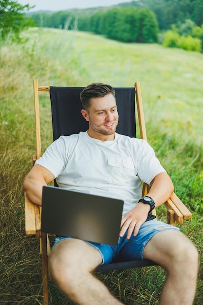 Een gelukkige man ontspant in de natuur en werkt op afstand op een draagbare laptop Werken op afstand terwijl hij op vakantie is Werken op een laptop terwijl hij in een stoel in de natuur zit