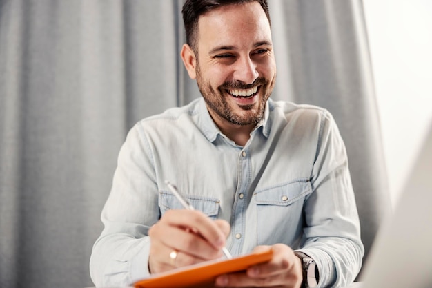 Een gelukkige ondernemer kijkt naar de laptop en schrijft in de agenda op zijn thuiskantoor