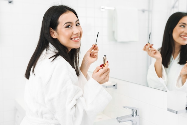 een gelukkige positieve jonge vrouw binnenshuis thuis in de badkamer past make-up toe.