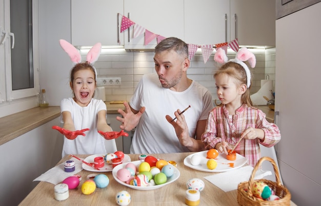 Een gelukkige vader met twee jonge dochters lacht en dobbert paaseieren schilderen en bereidt zich voor op de paasvakantie Voorbereiding voor de paasvakantie