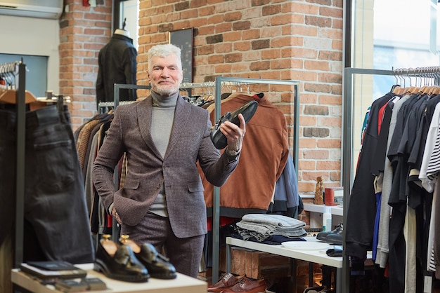 Een gelukkige volwassen man met grijs haar en een sportieve lichaamsbouw poseert met een zwarte schoen in een kledingwinkel. Een mannelijke klant met een baard draagt een wollen pak in een boetiek