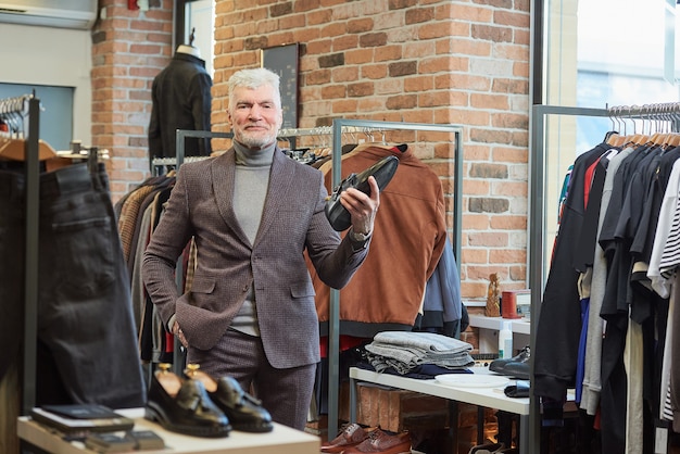 Een gelukkige volwassen man met grijs haar en een sportieve lichaamsbouw poseert met een zwarte schoen in een kledingwinkel. een mannelijke klant met een baard draagt een wollen pak in een boetiek