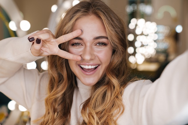 een gelukkige vrolijke jonge mooie vrouw neemt een selfie met de camera in de salon met vredesgebaar.