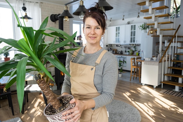 Een gelukkige vrouw in een groen huis met een potplant in haar handen glimlacht en zorgt voor een bloem Het interieur van een gezellig, milieuvriendelijk huis een open haardkachel een hobby voor het kweken en kweken van kamerplanten