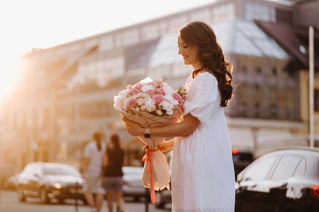Een gelukkige vrouw in een witte jurk bij zonsondergang met een boeket bloemen in de stad