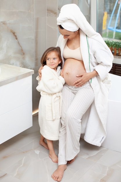 Een gelukkige zwangere moeder en oudste dochter in witte jassen knuffelen 's ochtends in de badkamer en kijken elkaar aan. Familie. Hoge kwaliteit foto