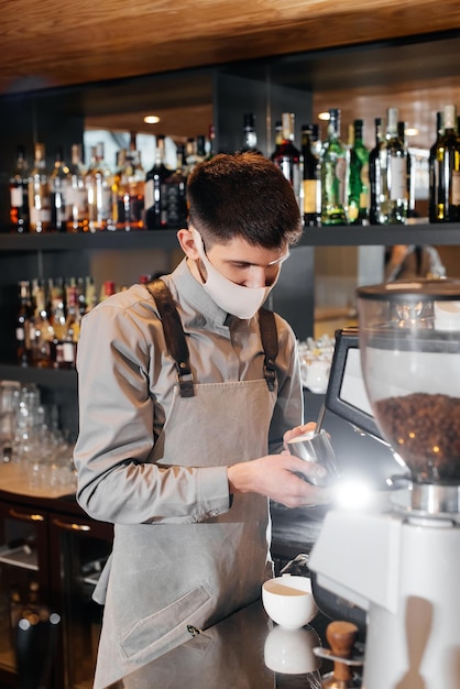 Een gemaskerde barista bereidt een voortreffelijke heerlijke koffie aan de bar in een coffeeshop Het werk van restaurants en cafés tijdens de pandemie