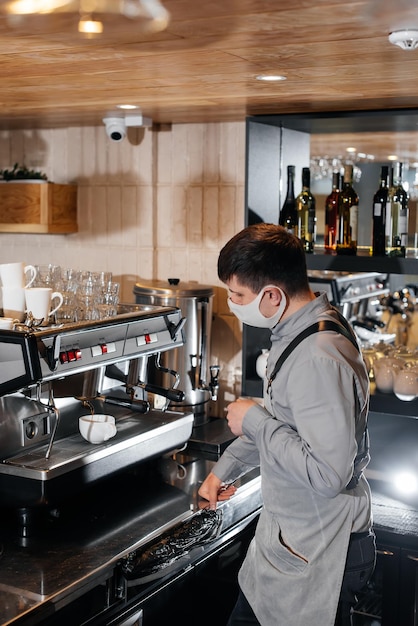 Een gemaskerde barista bereidt een voortreffelijke heerlijke koffie aan de bar in een coffeeshop Het werk van restaurants en cafés tijdens de pandemie